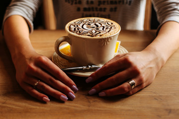 Fototapeta na wymiar The girl is drinking. The cup of coffee on a background of hands. Girl in a striped sweater with a cup of coffee. A cup of coffee with foam. Delicious patterns on the foam of coffee