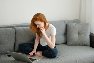 A red-haired girl in home clothes sits half-turn on the couch, wearily bending over a laptop