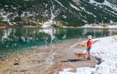 Little boy dressed a light red shirt walking by the mountain lake coast with his beagle dog friend. They enjoying spring atmosphere and melting snow landscape. Kids and pets friendship concept image.