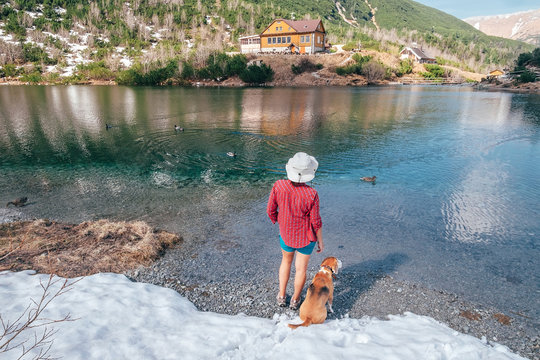 Little boy dressed a red light shirt walking by the mountain lake coast with his beagle dog friend. They enjoying spring atmosphere and melting snow landscape. Kids and pets friendship concept image.