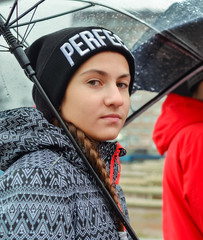 Lifestyle portrait of teenager girl under transparant umbrella wearing black knitted hat and casual rain jacket at rainy day in the city