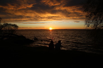 Two people at sunset with a camera sitting on the shore of a raging lake under storm clouds admiring the falling sun from a clear horizon.The fire is burning in half the sky.Russia