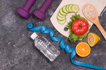 bottle of water, dumbbells, measuring tape, diet food on a black background, copy space, vegetables, fruits, oatmeal on a wooden spoon, weight loss and healthy lifestyle concept