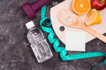 bottle of water, dumbbells, measuring tape, diet food on a black background, sticker for your text, vegetables, fruits, oatmeal on a wooden spoon, healthy lifestyle concept