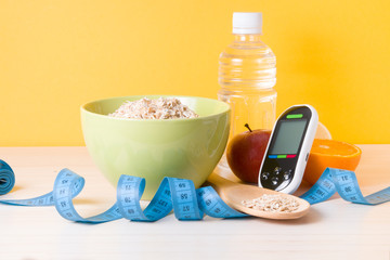 glucose meter, bottle of water, fruits, a bowl of oatmeal and a blue measuring tape on the table, yellow background, diabetes and blood sugar control concept, healthy lifestyle