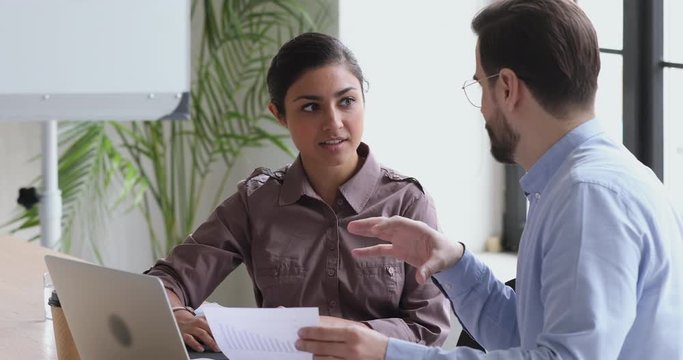 Focused young indian female trainee intern listening to skilled confident male boss leader supervisor, helping with paper documents, analyzing marketing data, giving professional advices at workplace.
