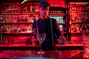 Professional barman preparing the alcoholic cocktail with shot in multicolored, deep red neon light, gives it to guest. Entertainment, drinks, service concept. Modern bar, trendy neoned colors.