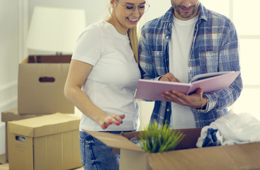 A happy middle-aged Caucasian husband and wife standing in their new home and taking out framed photo album out of cardboard box, chatting animatedly and laughing