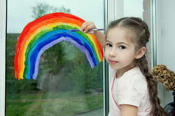Little smiling girl sits by the window on the windowsill and draws a rainbow on the glass