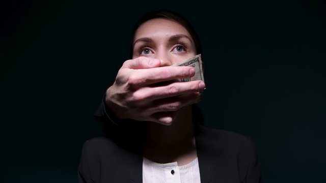 Money buys silence. Someone is covering a woman's mouth with dollars. Dark background, hand of an unknown buys the silence of a business woman in a jacket