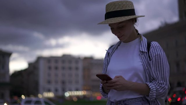 Caucasian female tourist in casual wear standing in urban setting and chatting in social networks on smartphone device in evening time.Young woman updating app on mobile phone using 4G internet

