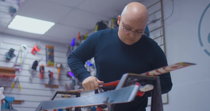 Professional craftsman with glasses removes an unnecessary layer from the surface of the skis with a scraper. Professional ski repair in the workshop, preparation for winter.