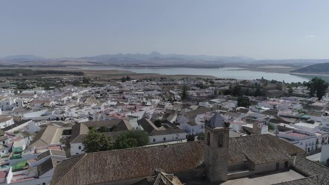 AERIAL DRONE ROOF TOP SHOT OF ANDALUSIAN TYPICAL WHITE SMALL VILLAGE. WITH CHURCH IN FIRST SHOT AND LAKE WITH MOUNTAIN RANGE IN HORIZON