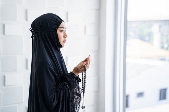 portrait side view of beautiful asian muslim woman looking up palms open to light praying to allah with prayer beads in the hands of the palm, wearing black hijab robe, in prayer room with cool tone