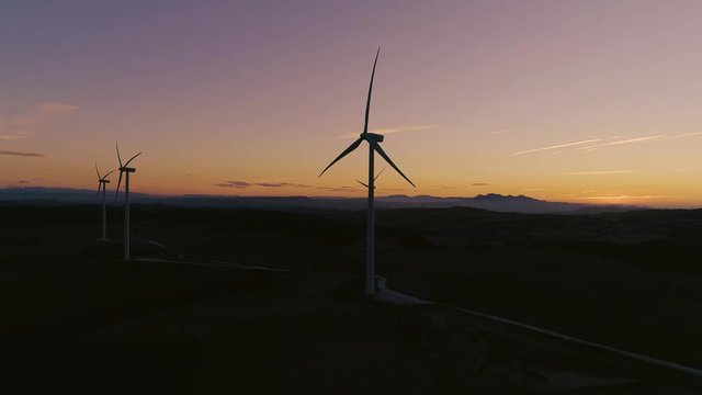 AERIAL DRONE SIDE SMOOTH IMAGE OF WIND TURBINES SPINNING WITH SUNSET AND MOUNTAIN RANGE 