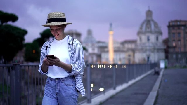Slow motion of happy young woman in casual wear dialing phone number on modern smartphone device and calling to friend using roaming connection during trip on weekends in evening time
