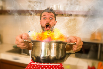 disaster home cook at kitchen- young funny and desperate man in cooking apron holding pot in flames in stress and fear making a mess of fire and smoke with food burning