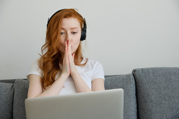 Red-haired girl in a white T-shirt and headphones sits on the couch, clasped palms to face and looks into the laptop