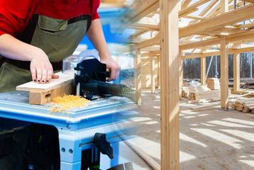 Woodworking. Concept - preparation of boards for construction. Human works with a grinding machine. Joiner aligns the boards. Concept - sale of lumber for construction. House frame closeup.