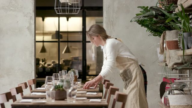 Young waitress preparing and laying the table in the restaurant. The girl sets plates and glasses.