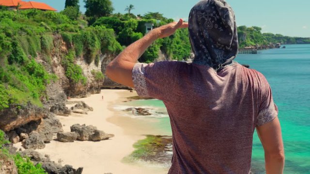 Traveler man on top of a mountain cliff takes photos and videos on smart phone beautiful tropical beach against the backdrop of the scenery nature landscape. Bali, Indonesia. 4K
