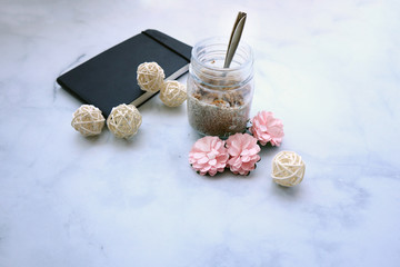 Fototapeta na wymiar A mason jar with chia pudding, pink flowers and a notebook on a marble surface with copy space
