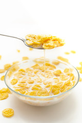 Side view of cornflakes drenched in milk in a spoon with which milk flows over a transparent bowl on a white background