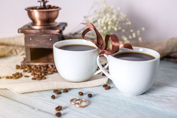 Couple of cups of fresh, aromatic black coffee are tied together with a ribbon. Wedding rings are placed in front of the mugs