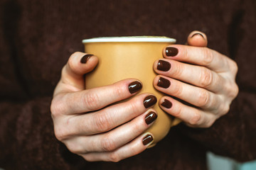 A woman wearing in a cozy sweater is holding a cup of aromatic coffee. The nails are coated with brown nail polish. Concept of elegance and stylish coffee manicure.