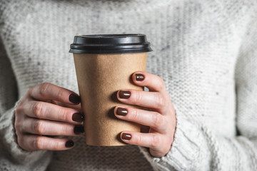 A woman wearing in a light cozy sweater is holding a paper cup of aromatic coffee. The nails are...