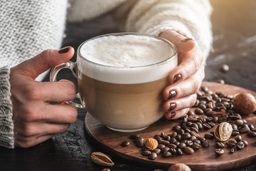 Women's hands in a warm sweater are holding a cup of fragrant latte. The nails are coated with brown nail polish. Coffee manicure. Breakfast or a break in a cozy cafe