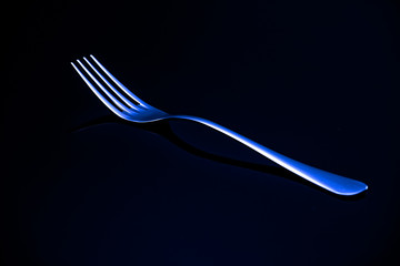 A metal fork with a blue neon reflection rests on a black glass. Advertising of the kitchen item