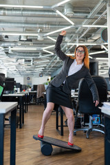 Cheerful business woman in red high heel shoes lost equilibrium on the balance board and falls. A female employee in a suit does sports at the workplace during the break. Bad coordination.