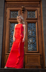 Sexy blonde with big blue eyes in a red evening dress poses gracefully on a city street