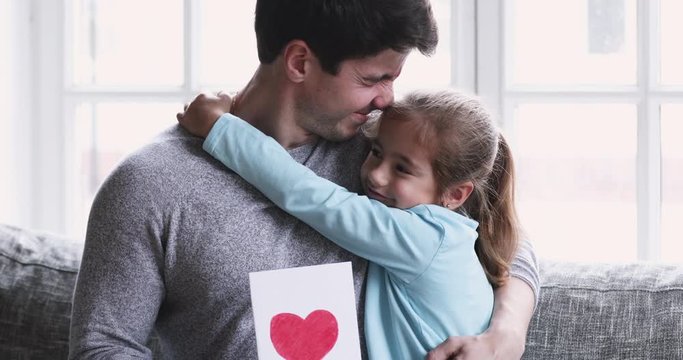 Happy young thankful father cuddling little adorable daughter, holding handmade card with drawn heart. Cute small child girl congratulating smiling daddy with Father's Day, presenting gift at home.