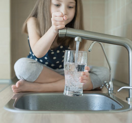Little girl open a water tap with her hand holding a transparent glass. Kitchen faucet. Glass of...