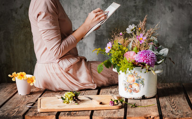 female artist draws a composition of wild flowers in a pot in a rustic style, a sketchbook on a...