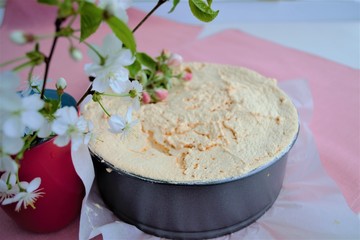 Homemade quark cake with meringue and caramel drops on top in a metal round baking sheet on baking paper and pink linen. On the left is a red glass with branches of flowering cherry and apple trees