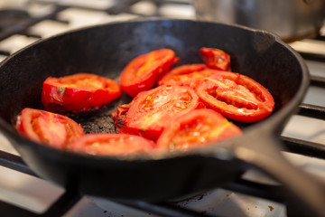 A closeup view of several Roma tomatoes cooking in a cast iron skillet pan, in a home kitchen setting.