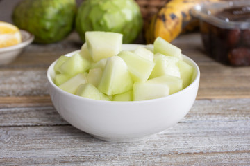 A view of a bowl of chopped honeydew melon pieces, in a still life setting.