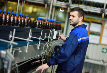 a young attractive male brewer or brewery worker in uniform against the background of a conveyor belt with plastic bottles of beer.