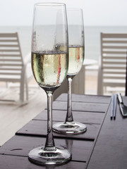 Two glasses of champagne of a wooden table near the sea, with the blur view of two white outdoor chaises in the background.