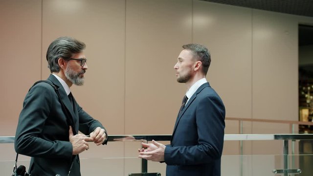 Business partners elegant men in formalwear are discussing work in office lobby standing together talking. Conversation and partnership concept.