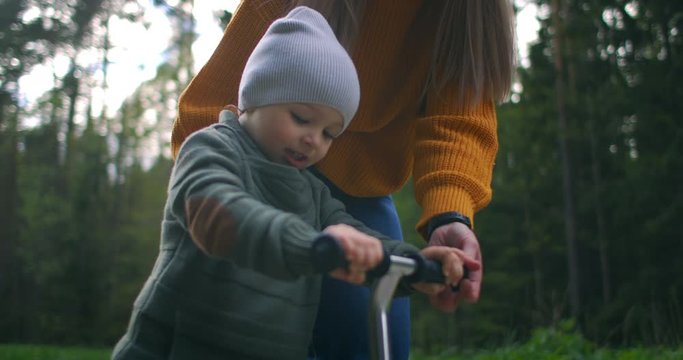  Mother and son drive scooter outdoors. He's always making us so proud. Cute little boy wearing cap riding mini scooter, kick scooter in the park in slow motion. Mom teaches to ride a scooter
