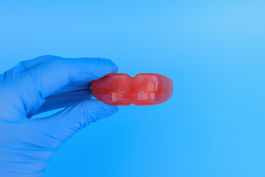 Doctor's hand in a blue glove holds a myofunctional trainer for teeth correction. Blue background. Selective focus.