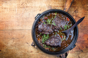 Traditional German braised beef cheeks in brown red wine sauce with carrots and onions offered as top view in a modern design stewpot on an old rustic board with copy space left