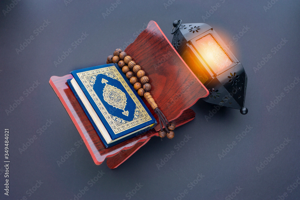 Wall mural Holy Al Quran with written arabic calligraphy meaning of Al Quran, lantern lamp and rosary beads or tasbih on black background.