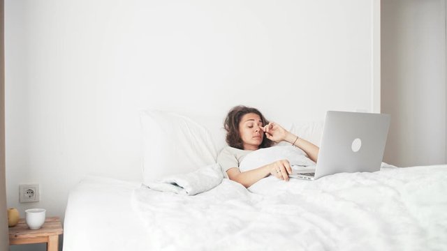 A young sleepy woman working at home using her laptop while lying and yawns in her bed in a bedroom.
