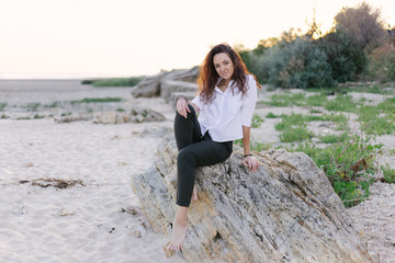 A woman sits on a wild beach,stones. freckles,dark curls hair,white shirt