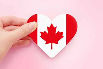 Female hand holds Canada heart shaped paper flag isolated over pink background, Canada Day...